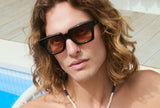 Man with curly hair wearing oversized black square frames with amber lenses by a sunlit pool