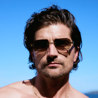 Shirtless man with wavy hair wearing aviator-style sunglasses with brown gradient lenses against blue sky