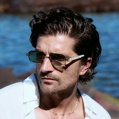 Man with wavy hair in white open-collar shirt wearing semi-rimless tortoise sunglasses by coastal water