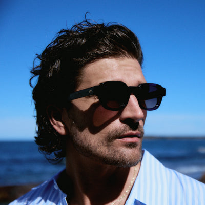 Man in blue striped shirt wearing chunky black square acetate sunglasses against an ocean horizon
