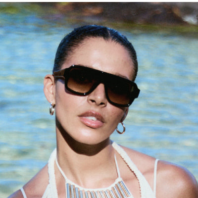 Sleek-haired woman in cream knit top wearing oversized shield-style sunglasses by rippled water